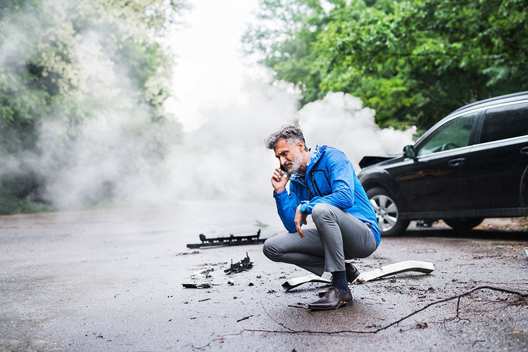 Mature man making a phone call after a car accident, smoke in the background.